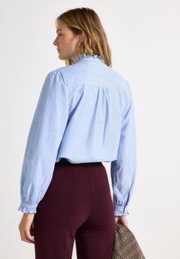 Hemelsblauwe blouse met lange mouwen, een gerimpelde kraag en een geplooide achterkant, gecombineerd met hoog getailleerde bordeauxrode broek en een gestructureerde jas.