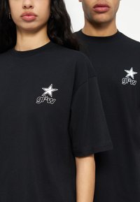 Δύο άτομα φορούν μαύρα μπλουζάκια με λευκό αστέρι και λογότυπο «gl*w» στο πάνω αριστερό μέρος του στήθους, σε απλό φόντο.