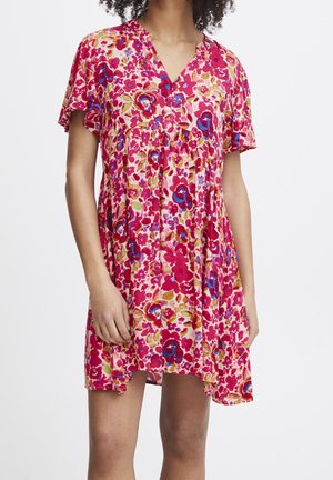 Frau trägt ein kurzärmeliges, V-Ausschnitt-Kleid mit einem lebendigen roten, pinken, blauen und gelben Blumenmuster, steht vor einem schlichten weißen Hintergrund.