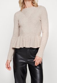 Beige geripptes Strickpullover mit langen Ärmeln, tailliertem Oberteil und ausgestellt Peplum-Saum. Getragen mit schwarzen Kunstlederhosen.