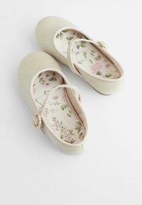 Bailarinas pálidas y brillantes con punta redonda, correa de color crema y plantilla con un patrón floral. Acento decorativo de hardware dorado cerca de la correa.