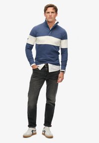 Mannen navyblauwe pullover met witte horizontale streep, ritskraag, gedragen met zwarte jeans en witte sneakers met grijze en zwarte accenten.