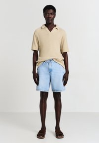 Junger Mann steht, trägt einen beigen, gestrickten Kurzarmpullover, hellblaue Jeansshorts mit Kordelzug und braune Sandalen vor einfarbigem Hintergrund.