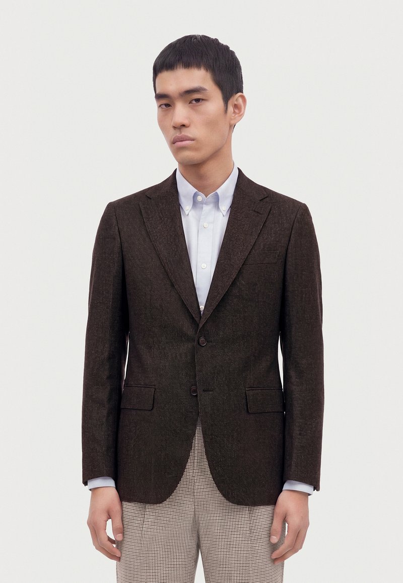 Homme portant un blazer marron foncé sur une chemise blanche à boutons avec un pantalon à carreaux clair, debout devant un fond clair uni.