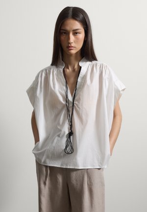 Mujer con cabello largo y oscuro que lleva una blusa blanca suelta, pantalones beige y un largo collar de cuentas negras, de pie con las manos en los bolsillos.