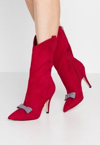 Bottes chelsea en suede rouge avec des bouts pointus, des talons fins et des accents de nœuds à carreaux sur les dessus; dotées de zips latéraux et de contours élégants.