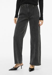 Jeans de mezclilla negros de cintura alta con corte de pierna ancha y detalles de costuras visibles, combinados con tacones negros puntiagudos.