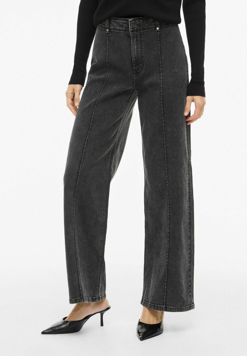 Jeans de mezclilla negros de cintura alta con corte de pierna ancha y detalles de costuras visibles, combinados con tacones negros puntiagudos.