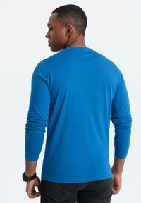 Man som bär en klarblå långärmad skjorta och svarta jeans, sedd bakifrån, stående mot en enkel vit bakgrund.