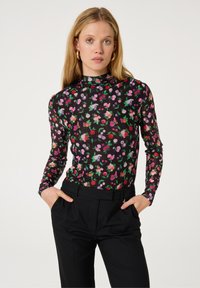 Langsleeve top in doorschijnende zwarte stof met een kleurrijk bloemenpatroon in roze, paars, rood en groen. Gecombineerd met hoge zwarte broeken.