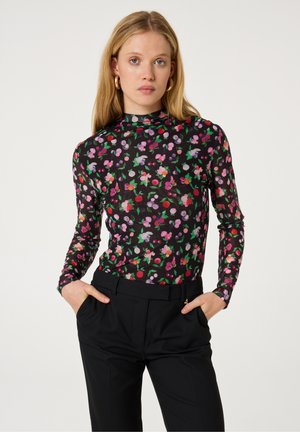 Langsleeve top in doorschijnende zwarte stof met een kleurrijk bloemenpatroon in roze, paars, rood en groen. Gecombineerd met hoge zwarte broeken.