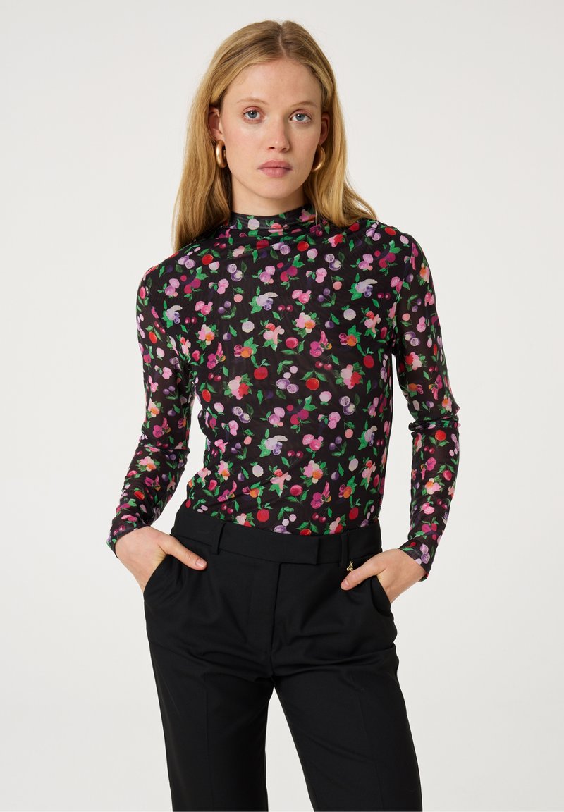 Langsleeve top in doorschijnende zwarte stof met een kleurrijk bloemenpatroon in roze, paars, rood en groen. Gecombineerd met hoge zwarte broeken.