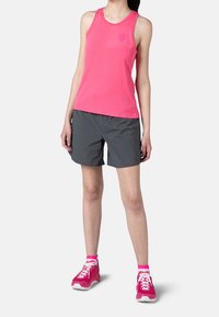 Žena stojící v růžovém topu bez rukávů, šedých šortkách, růžových ponožkách a růžových teniskách na bílém pozadí.