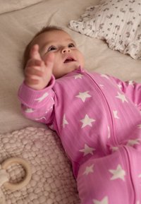 Grenouillère bébé rose avec motifs d'étoiles blanches, en tissu texturé, dotée d'une fermeture éclair à l'avant. Couverture en peluche et coussin floral en arrière-plan.