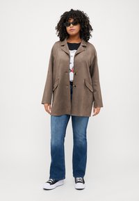 Brown suede jacket with a collared neckline, two front pockets, and button closure, paired with blue jeans and black sneakers.