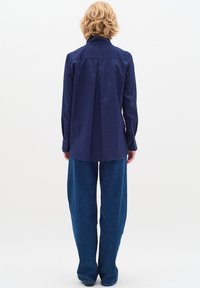 Persona con capelli biondi corti, che indossa una camicia a maniche lunghe blu navy e jeans azzurri larghi, in piedi con la schiena rivolta verso un sfondo bianco.