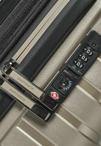 Valigia beige chiaro con zip nere. Dotata di lucchetto a combinazione TSA, hardware argentato e superficie testurizzata. Gli accenti includono dettagli di branding.