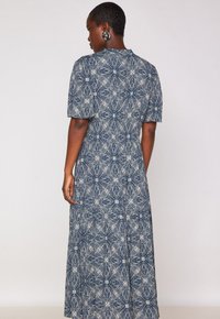 Femme aux cheveux courts portant de grandes boucles d'oreilles noir et blanc et une longue robe à motif bleu marine et blanc, de dos contre un fond uni.