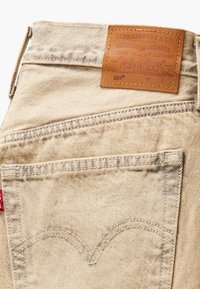 Béžové džínsy z denimu s koženou nášivkou značky, s kontrastným prešívaním a charakteristickým dizajnom zadnej vrecka. Štýl 501, štandardný strih.