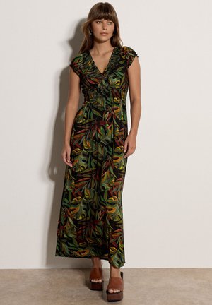 Frau steht und trägt ein langes, kurzärmeliges schwarzes Kleid mit grünen, gelben und roten Blattmustern und braune Plateausandalen.