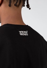 Černé bavlněné tričko s kulatým výstřihem a bílým nápisem "WRONG FRIENDS" vytištěným na horní části zad. Hladký povrch.