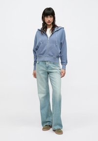 Ανοιχτό μπλε φούτερ με φερμουάρ και άνετη εφαρμογή, σε συνδυασμό με ανοιχτόχρωμο, καμπανιστό τζιν και πράσινο-καφέ παπούτσια. Απλός, καθημερινός σχεδιασμός.