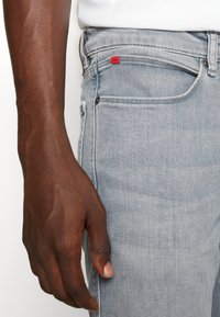 Light blue denim jeans with a fitted design, featuring a red brand tag and metal button detail, held by a hand against a plain background.