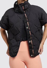 Femme portant une veste matelassée noire à manches courtes avec boutons pression et des leggings roses, mains posées sur les hanches sur un fond uni.