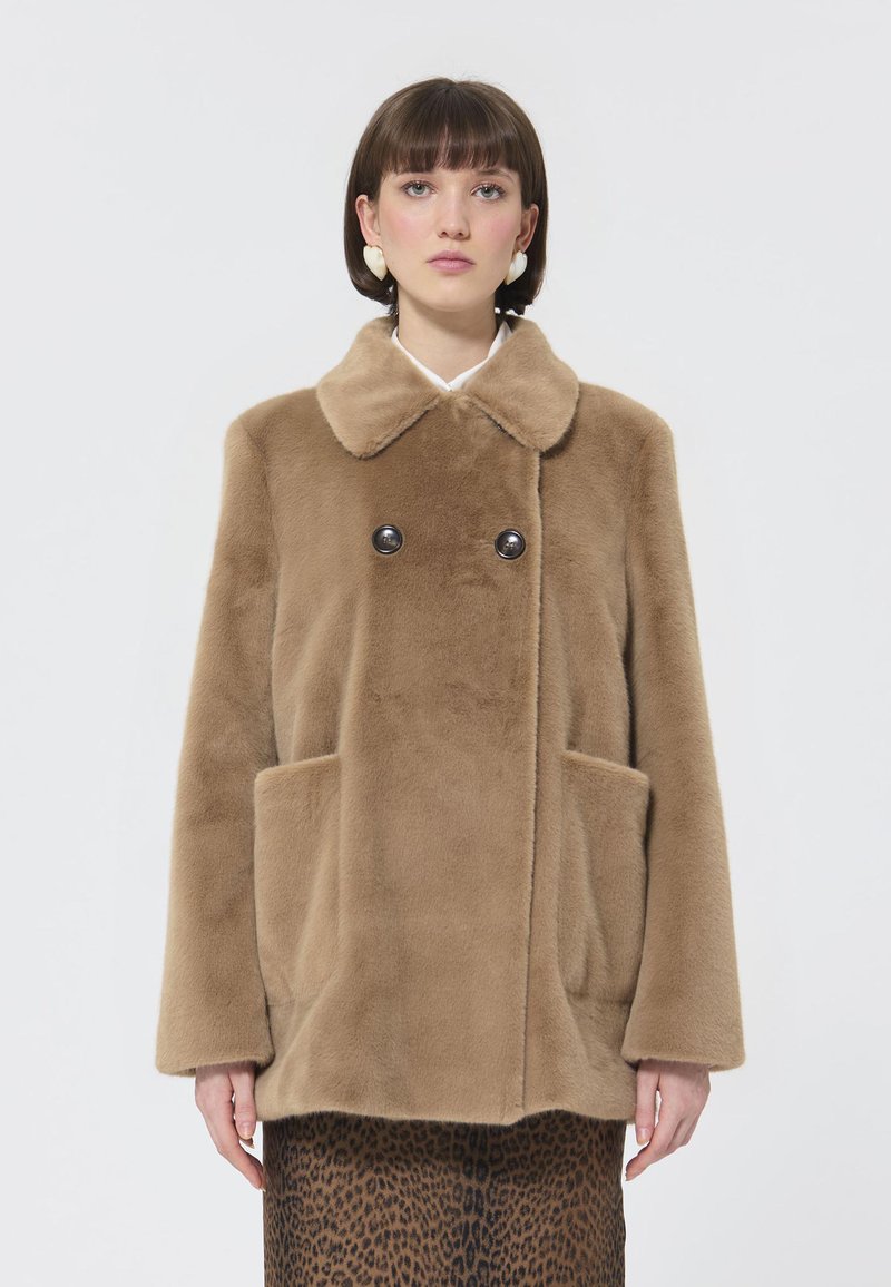 Cappotto in ecopelle marrone con ampio colletto, chiusura a doppio bottone e tasche laterali, indossato sopra una gonna con stampa leopardata. La morbidezza della texture è predominante.