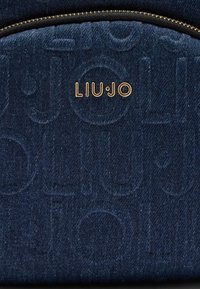 Tamno plava traper ranac s urezanim "LIU JO" logotipom. Ima zlatni zatvarač i teksturiranu površinu s suptilnim uzorcima.