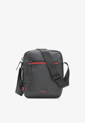 Kompaktowa czarna torebka na ramię z regulowanym paskiem, przednia kieszeń na zamek z czerwonym akcentem oraz czerwonym logo marki na dole, na środku.