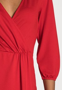 Blouse cache-cœur rouge avec col en V, manches trois-quarts, taille froncée et texture douce. Caractérisée par un tissu lisse et léger.