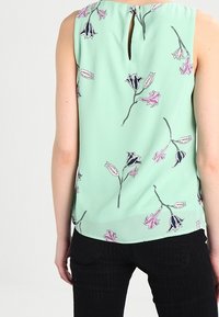 Blusa senza maniche verde menta con motivo floreale in rosa e nero. Presenta un dettaglio a chiave sulla schiena e tessuto leggero e liscio.