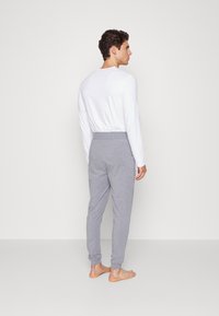 Grå joggebukser med elastiske mansjetter, laget av mykt materiale, sammen med en hvit langermet topp. Modellen står barbeint, vendt bort.