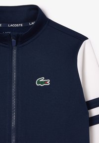 Granatowa kurtka zapinana na zamek z kontrastowymi białymi rękawami. Posiada teksturowany materiał, pasek akcentowy oraz zielone logo krokodyla na klatce piersiowej.