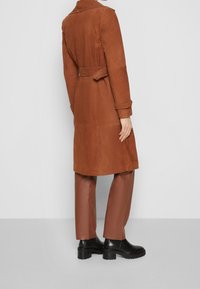 Gabardina de ante marrón con cinturón en la cintura, mangas largas y cuello con solapas, combinada con pantalones marrones de cuero sintético y botines negros.