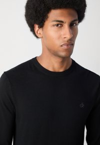 Black long-sleeve sweater made of soft fabric featuring a round neck and a small logo on the chest. Simple, fitted design.