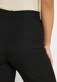 Černé elegantní kalhoty s hladkou texturou, úzkým střihem a jednou zadní kapsou s horizontálním klopou.