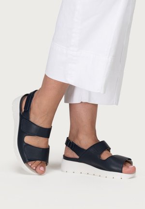 Füße, die marineblaue Sandalen mit offenen Zehen, weißen Plateausohlen und verstellbaren Riemen tragen, kombiniert mit weißen, gekürzten Hosen.