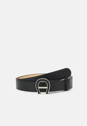 Ceinture en cuir noir texturé avec une boucle argentée en forme de fer à cheval, roulée sur un fond clair uni.
