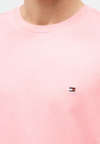 Κοντινό πλάνο ενός ατόμου που φορά ένα ανοιχτό ροζ T-shirt με στρογγυλή λαιμόκοψη και ένα μικρό ορθογώνιο λογότυπο σε κόκκινο, λευκό και ναυτικό μπλε στο στήθος.