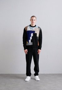 Pull en tricot avec des motifs abstraits multicolores bleu, brun et noir. Assorti à un pantalon noir et des baskets blanches.