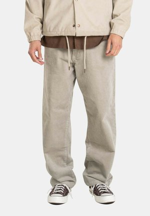 Mann trägt beige Cordhosen, dunkelbraune Sneakers mit weißen Schnürsenkeln und eine beige Jacke über einem braunen Hemd.