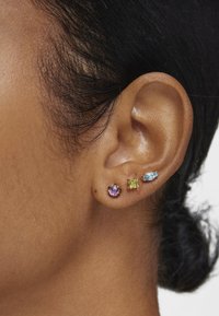 Tres pendientes de stud en varias formas: una flor morada, un cuadrado verde y una forma marquise azul, en metal dorado. El tono de piel es cálido.