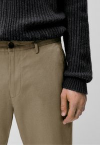 Zwarte geribbelde trui met een hoge halslijn, gecombineerd met beige broek met een knoopsluiting en subtiele zijzakken, waarbij handdetails zijn te zien.