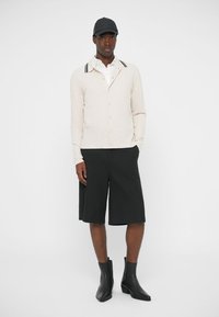 Bēšas krāsas krekls ar pogām un melnas krāsas apkakles svītrām, kombinēts ar melniem ceļgala garuma šortiem un melnām potīšu zābakiem uz vienkrāsaina fona.