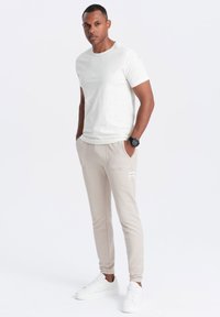 Camiseta blanca de manga corta, joggers beige con bolsillos laterales y zapatillas blancas; diseño casual, textura suave, detalles de marca mínimos.