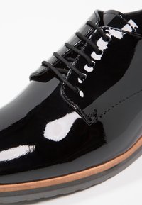 Chaussure habillée en cuir verni noir avec une finition lisse, bout rond, six œillets et lacets noirs ; équipée d'une semelle en cuir marron.