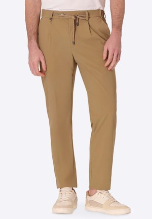 Uomo che indossa pantaloni beige a gamba stretta con coulisse in vita e sneakers beige casual, in piedi contro uno sfondo semplice.