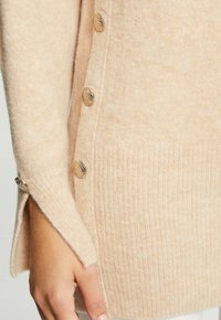 Pull beige en maille avec texture côtelée, comprenant des boutons dorés le long du côté et un ourlet légèrement allongé avec une fente latérale.
